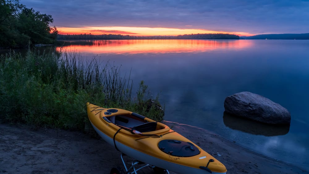 A kayak on a sandy shore with a calm lake and a sunset in the background.