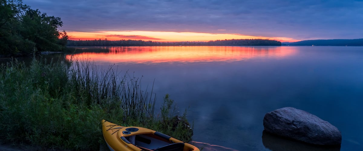 A kayak on a sandy shore with a calm lake and a sunset in the background.