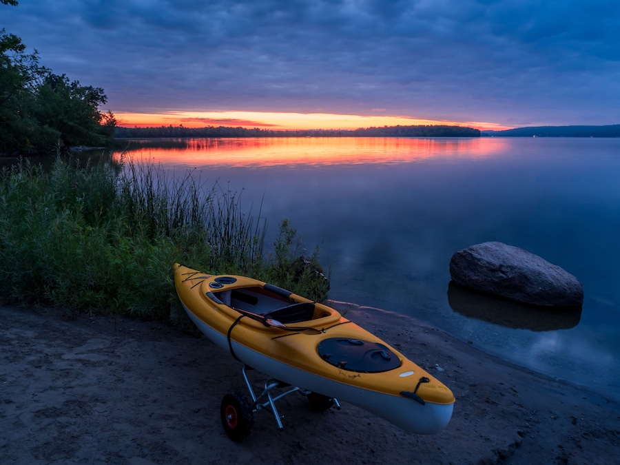 A kayak on a sandy shore with a calm lake and a sunset in the background.