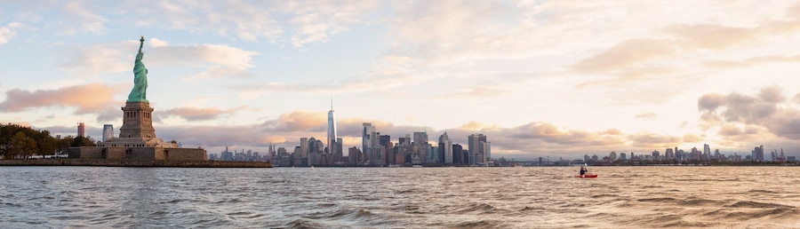 Panoramic view of the Statue of Liberty and Downtown Manhattan in the background during a vibrant cloudy sunrise. Taken in Jersey City, New Jersey, United States.
