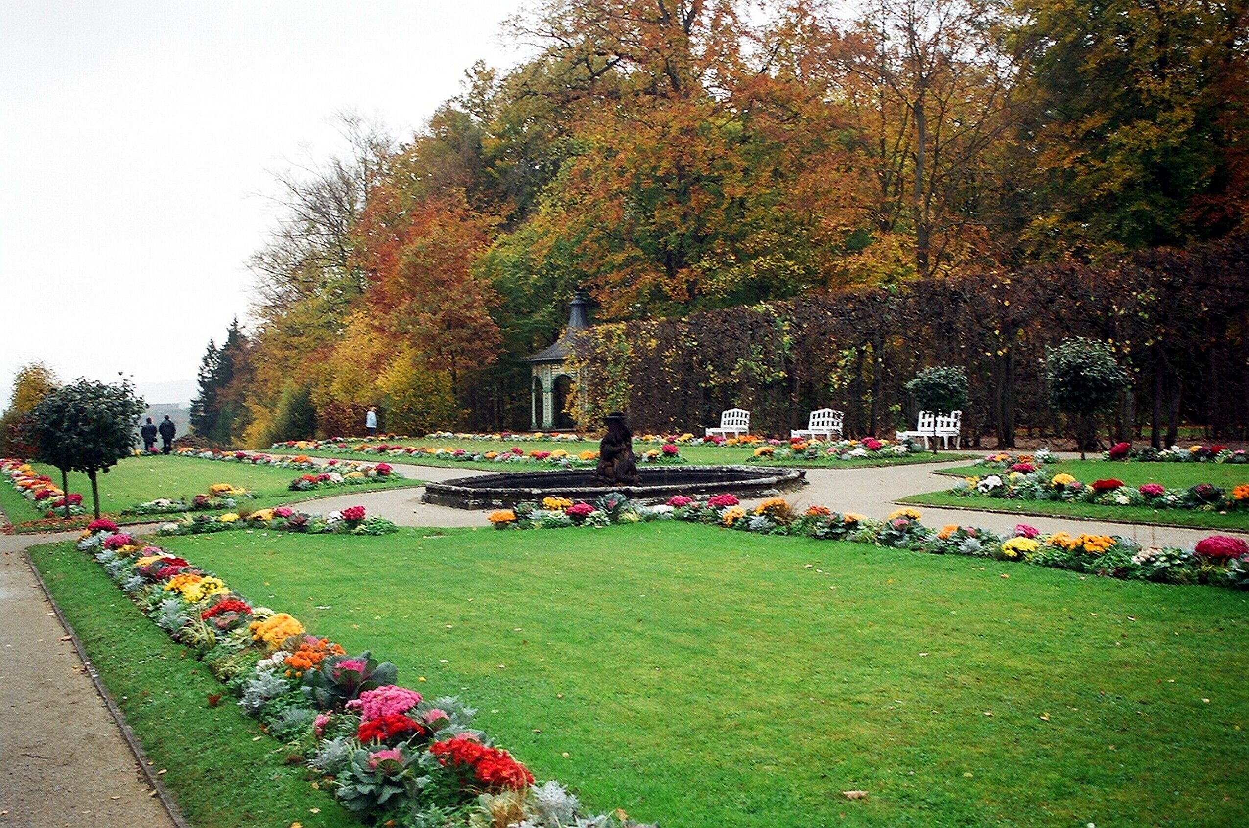 Um parque com uma fonte central, rodeado por canteiros de flores e árvores com folhagem outonal.