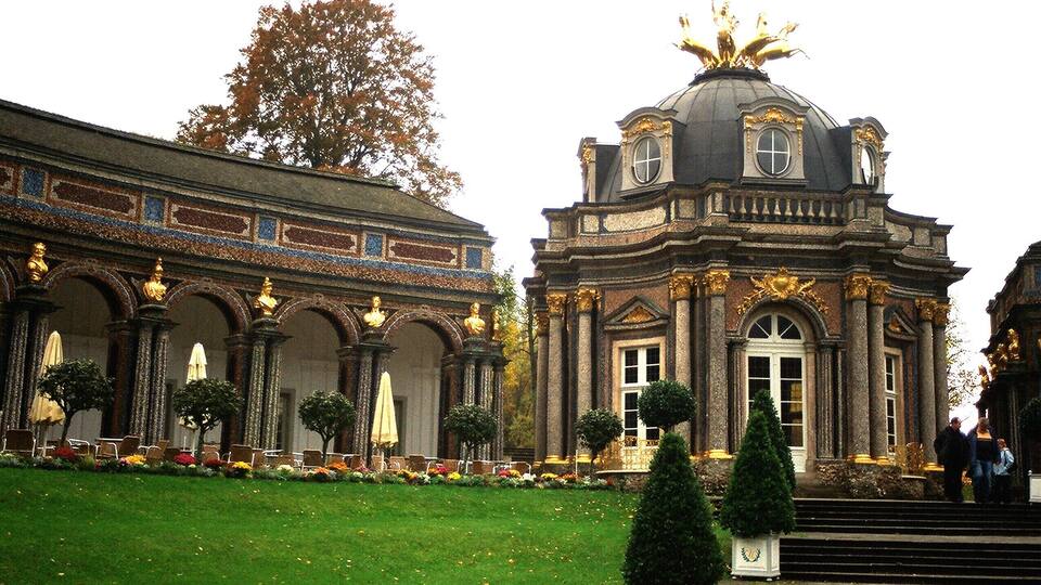 A grand building with ornate architecture, including a dome and columns, surrounded by a well-maintained garden with trees and flowers.