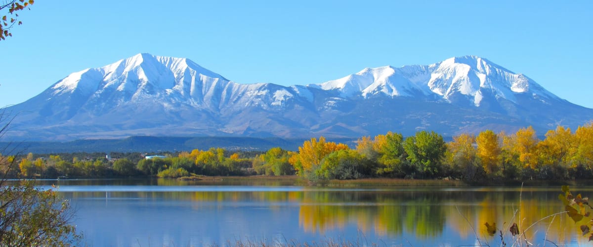 A mountain range with snow-capped peaks, a calm lake, and autumn-colored trees.