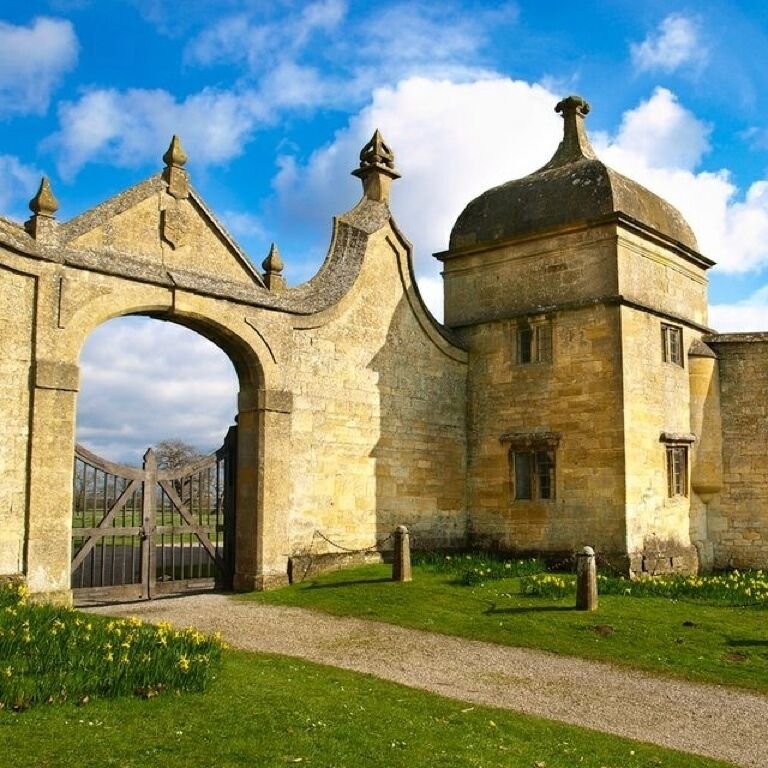 A historic stone gate with a large archway, a small tower, and a grassy area with yellow flowers.