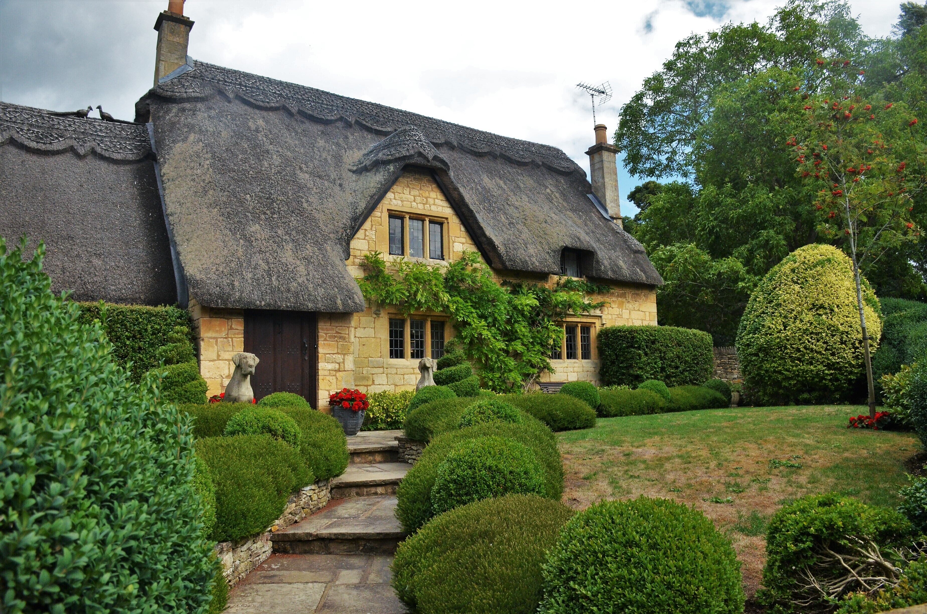 A thatched-roof cottage with a stone facade, surrounded by well-manicured hedges and a small garden.