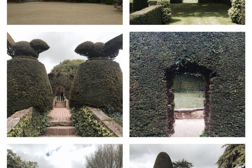 Um edifício histórico de pedra com árvores em flor, uma grande árvore em um parque, arte em topiaria, um arco de sebes, um caminho de sebes bem aparado e um jardim com tulipas.