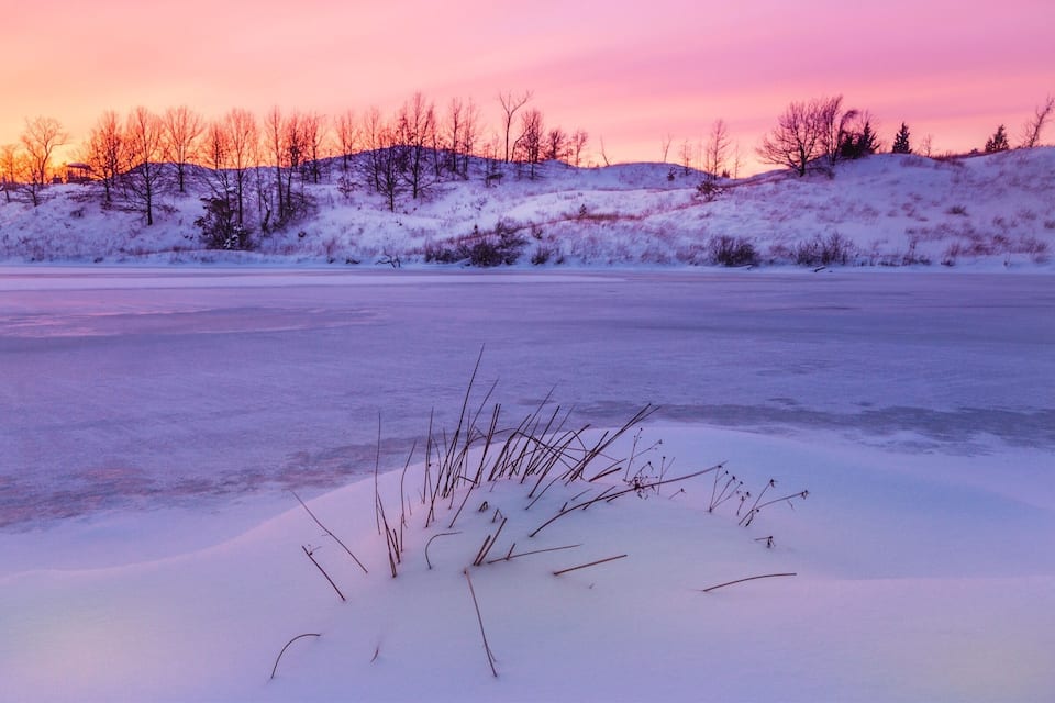 Un lago helado con poca vegetación en primer plano y un cielo al atardecer.