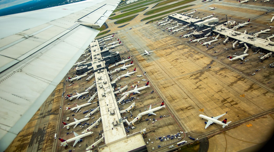 Una pista de aeropuerto con múltiples aviones estacionados en las puertas de embarque, una torre de control y un paisaje urbano distante.