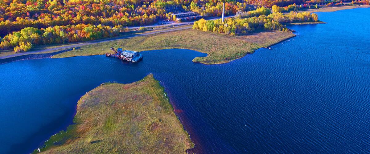 An aerial view of a river with a boat, surrounded by autumn-colored trees and a small island with buildings.