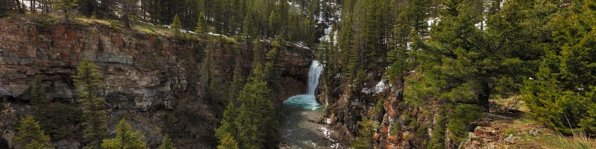 A canyon with a waterfall, dense forest, and a snow-capped mountain.