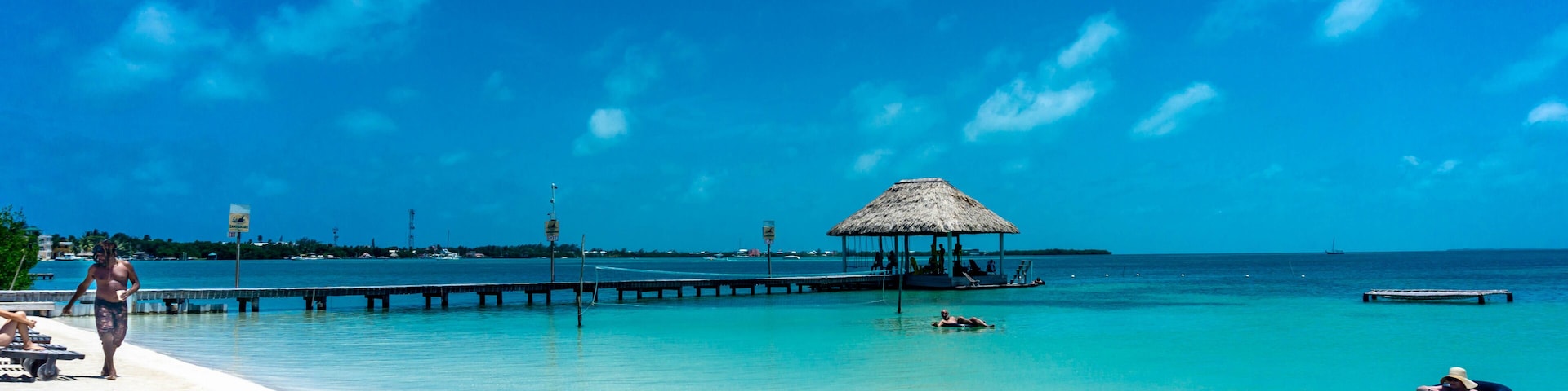 A beach with clear turquoise water, a thatched-roof hut, a pier extending into the sea, and a person floating on a raft.