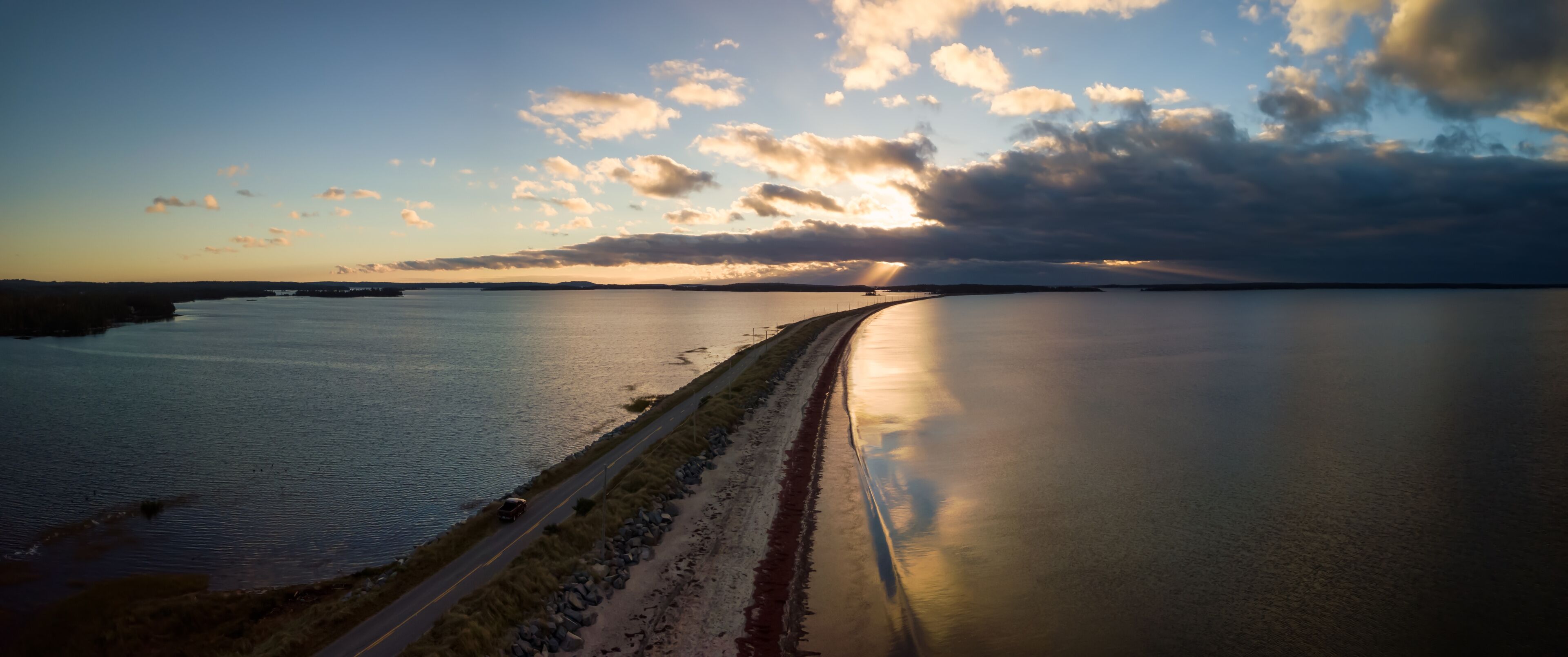 A coastal road curving along a calm body of water with a sunset in the background.