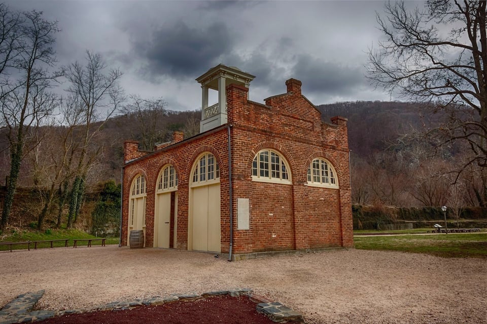A brick building with arched windows and a chimney, surrounded by a gravel area and trees.