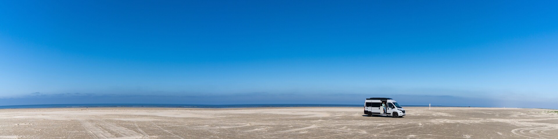 panorama of a gray camper van parked on an endless white sand beach in the middle of nowhere with ocean behind