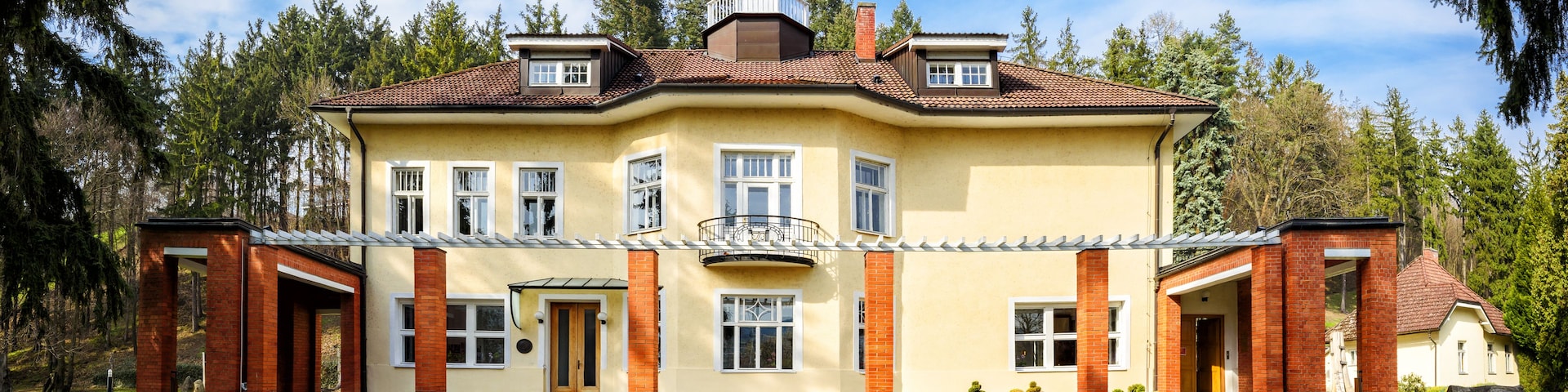 A large, two-story house with a red-tiled roof, surrounded by a well-maintained lawn and tall trees.