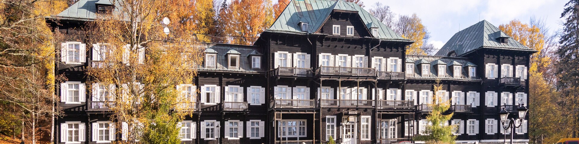 Un edificio grande de varios pisos con fachada oscura y contraventanas blancas, rodeado de árboles de colores otoñales y un camino adoquinado.