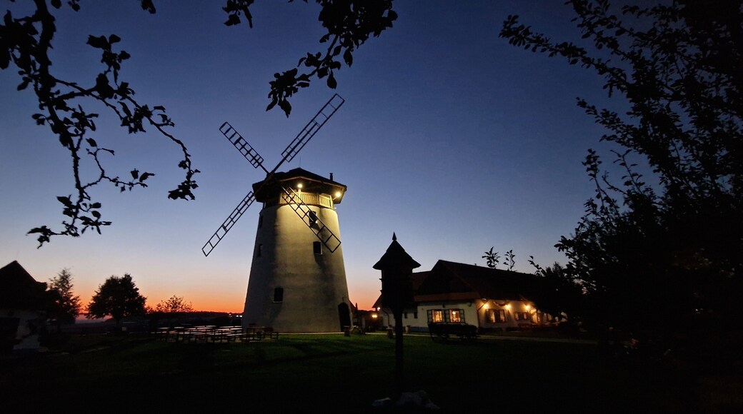A windmill at dusk with a lit top, surrounded by trees and a small building.