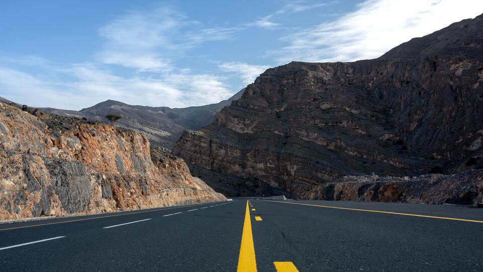 A road with yellow center lines, flanked by rocky cliffs and mountains under a clear sky.