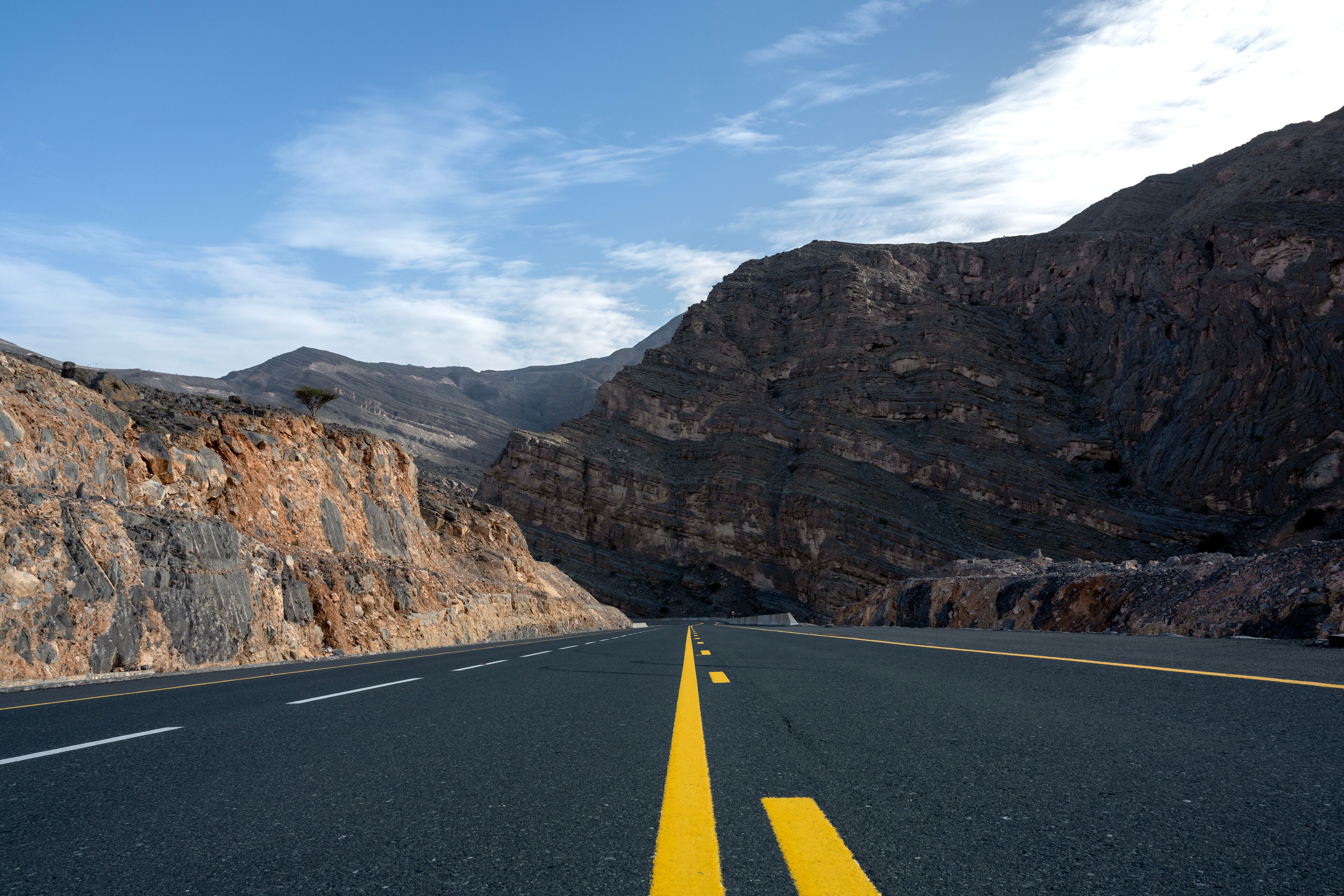 A road with yellow center lines, flanked by rocky cliffs and mountains under a clear sky.
