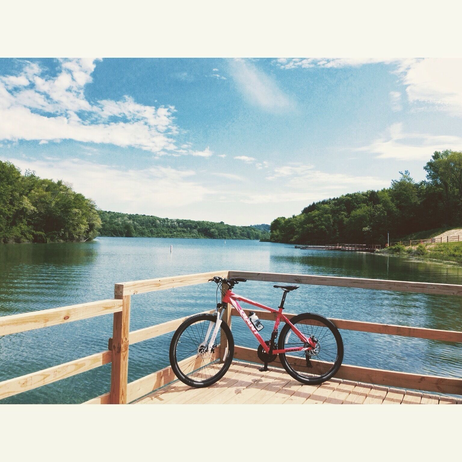 A red bicycle parked on a wooden dock by a calm lake with a wooden railing.