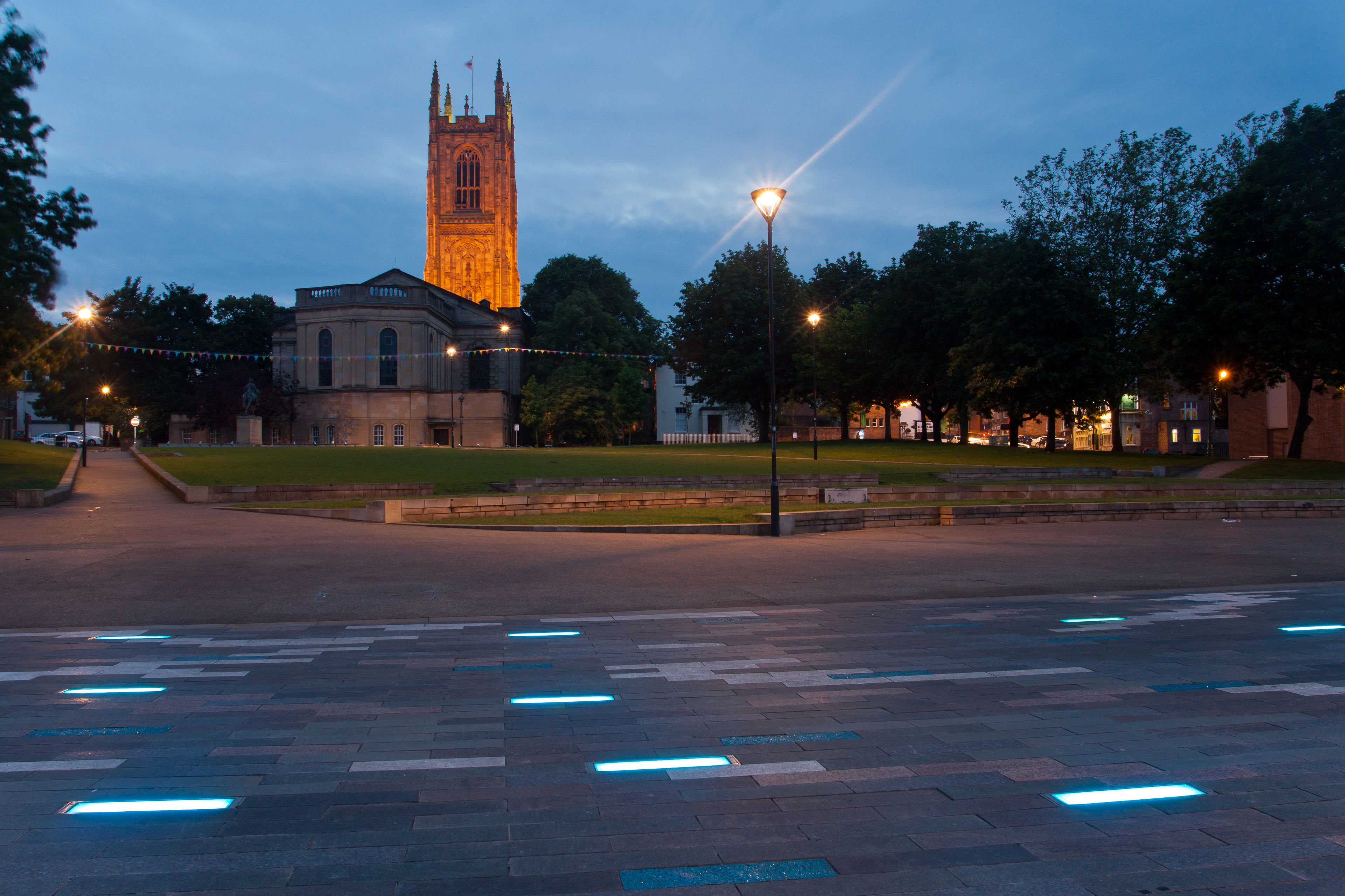 A historic building with a tall tower, a paved plaza with glowing lines, and trees in the background.