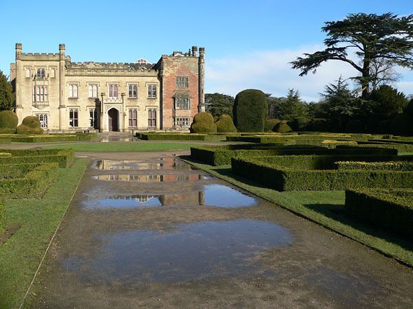 A historic building with a symmetrical facade, a central arched entrance, and a formal garden with hedges and a reflecting pond.