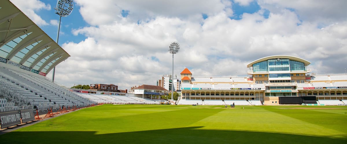 A large, empty stadium with a green field, a tall floodlight, and a modern building with a clock tower.