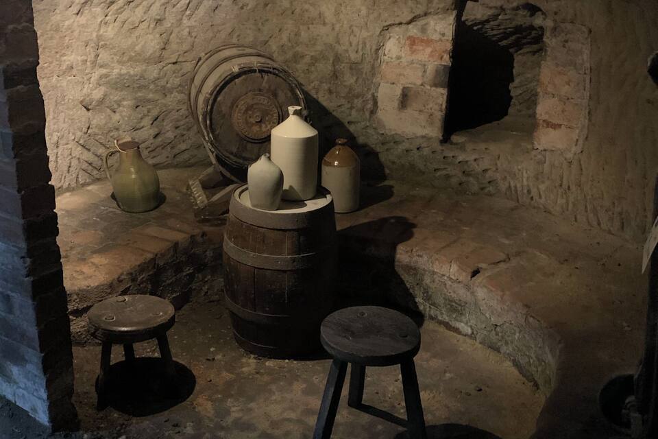 A rustic interior with stone walls, a wooden barrel, and several ceramic containers.