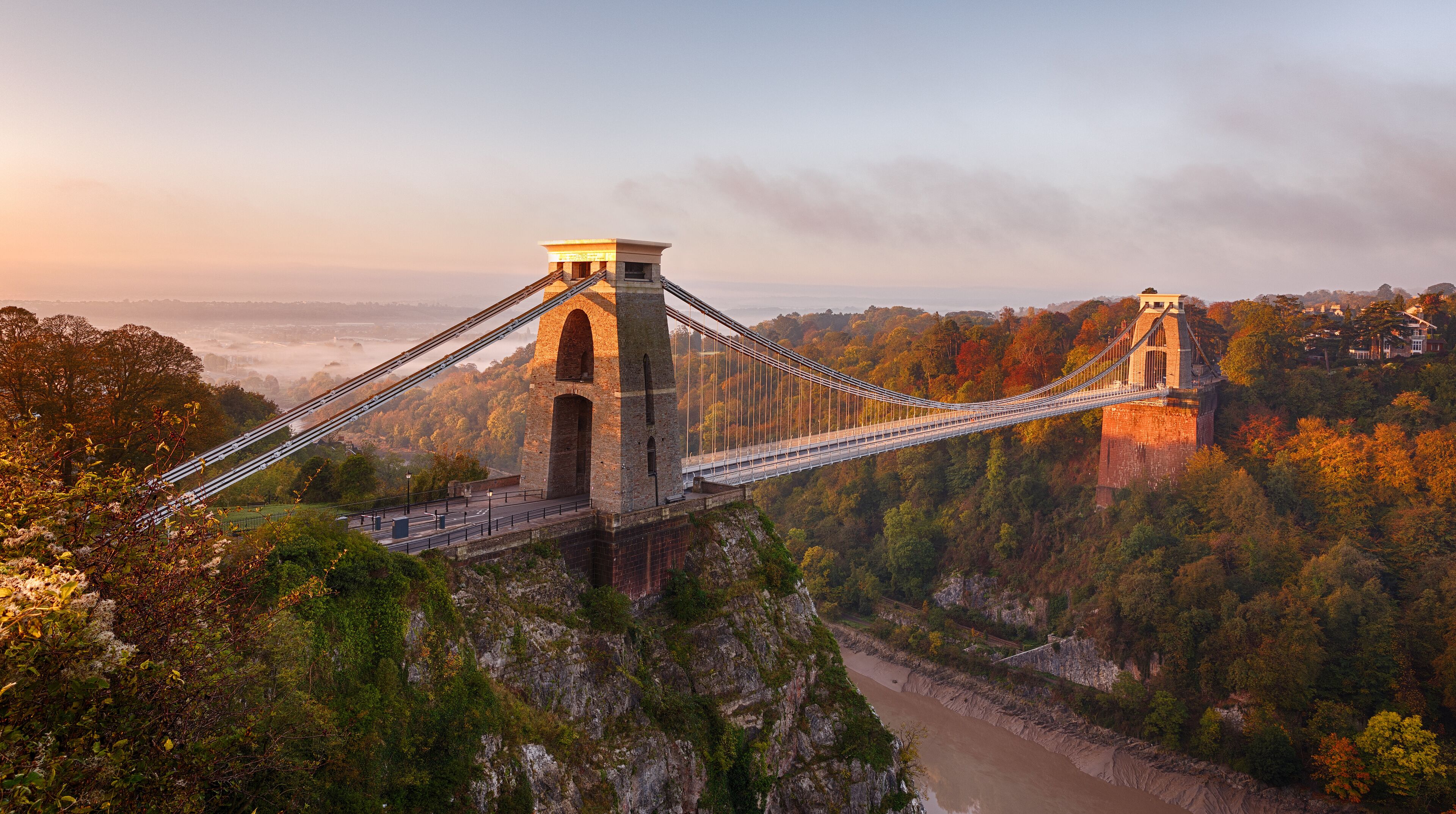 A suspension bridge with two towers, surrounded by autumn foliage and a river below.