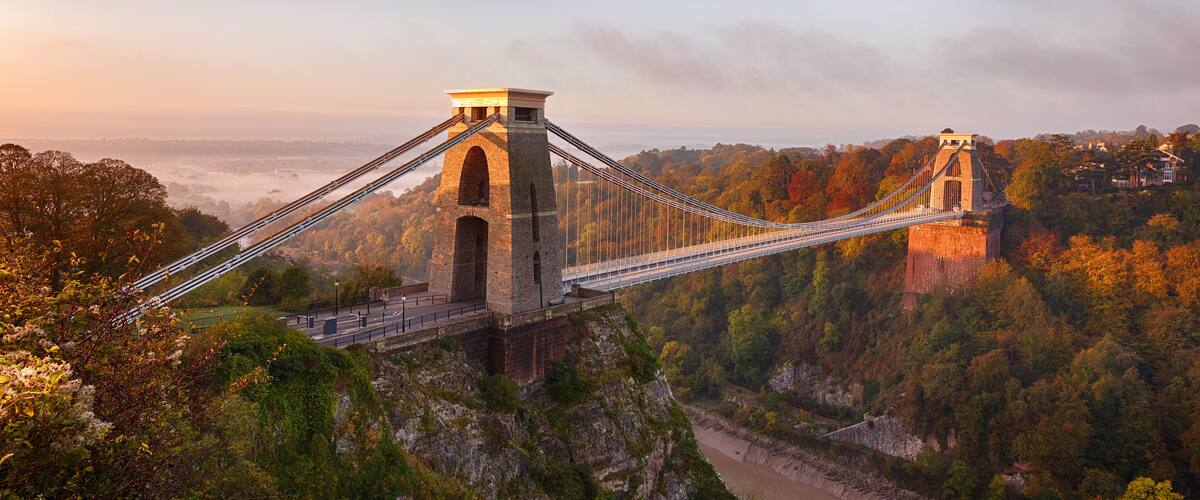 A suspension bridge with two towers, surrounded by autumn foliage and a river below.