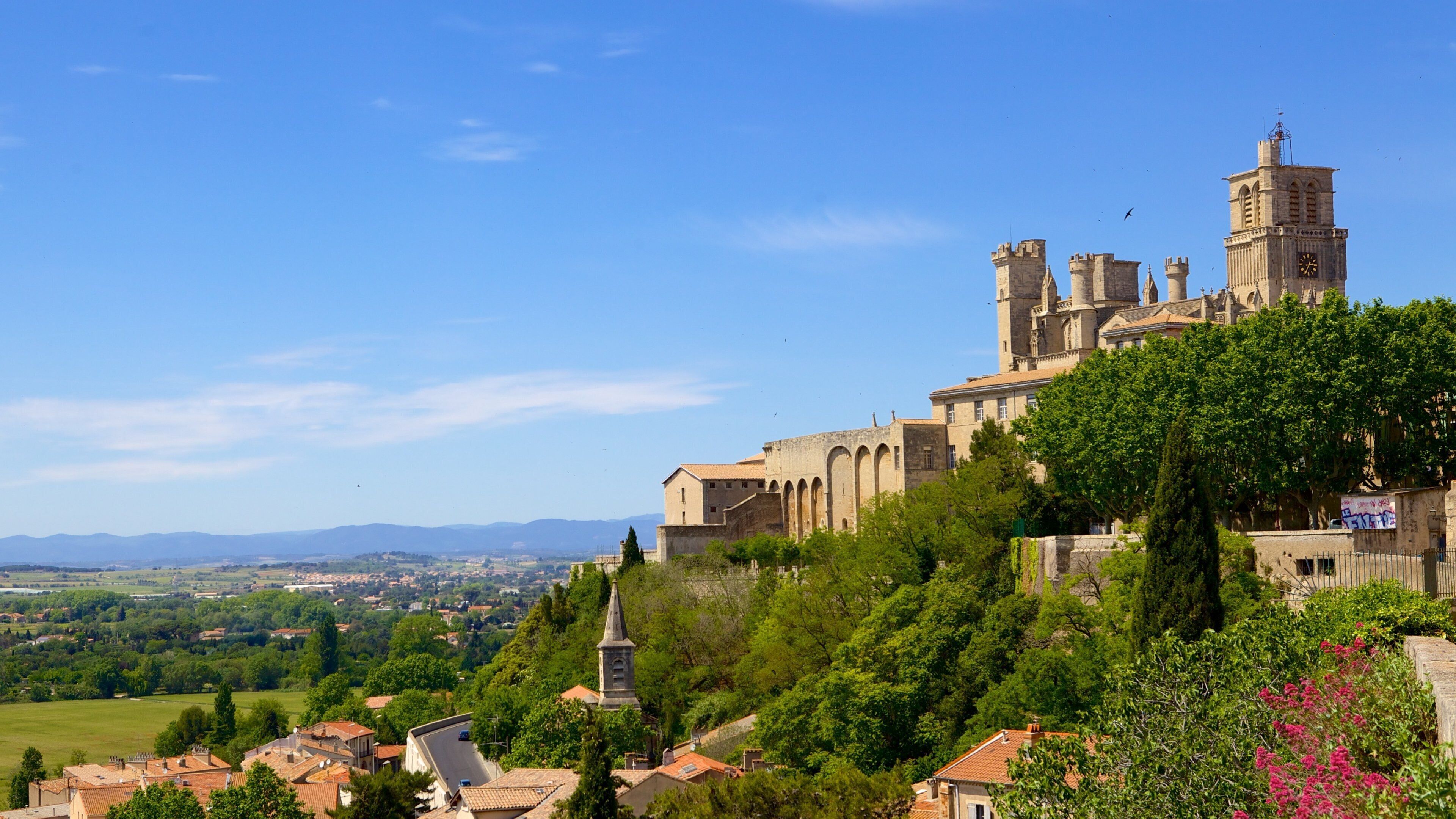 A historic castle with towers and a clock tower, surrounded by lush greenery and a distant view of the countryside.