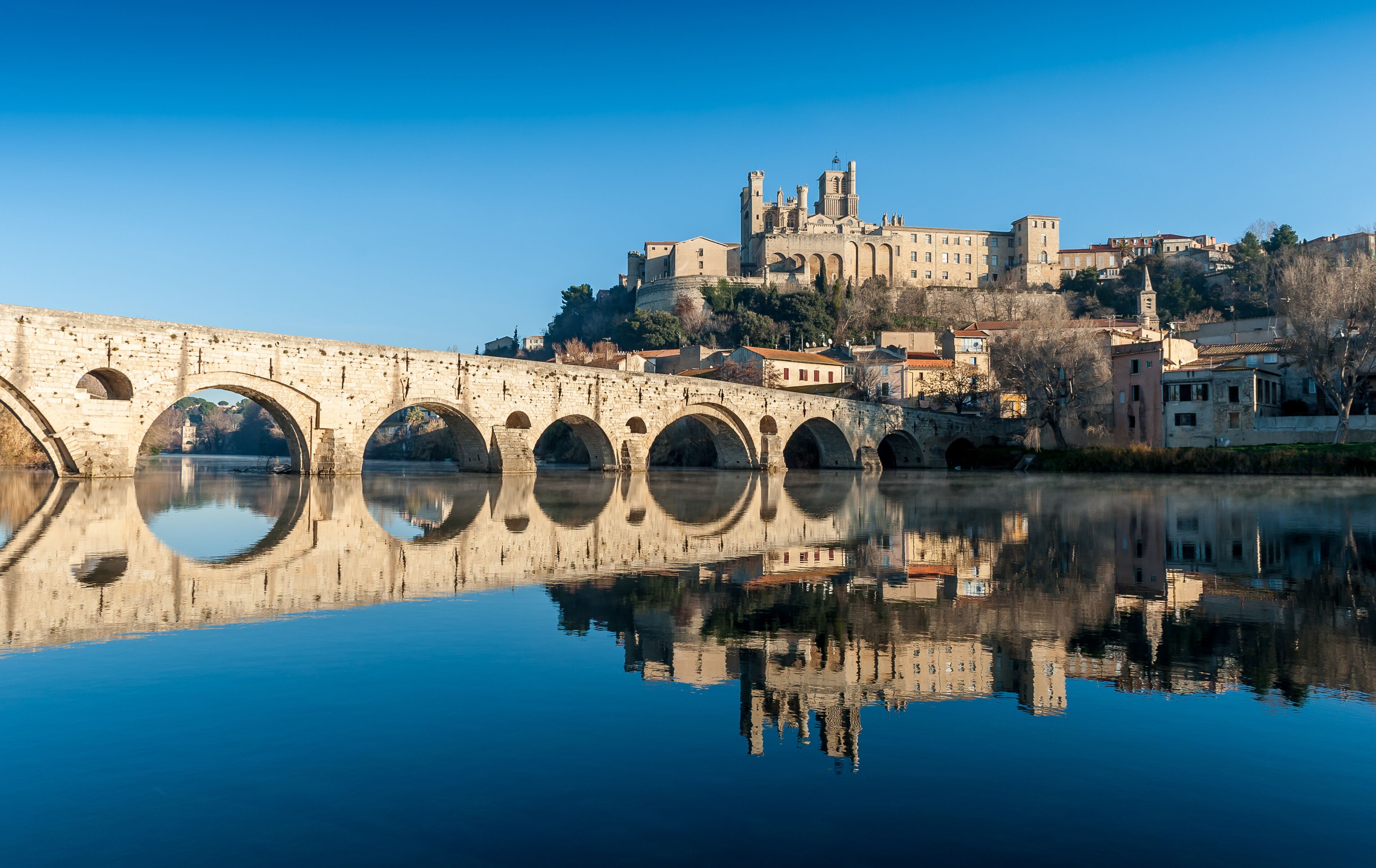 A stone bridge with arches reflecting in calm water, a historic building with towers in the background, and a clear blue sky.