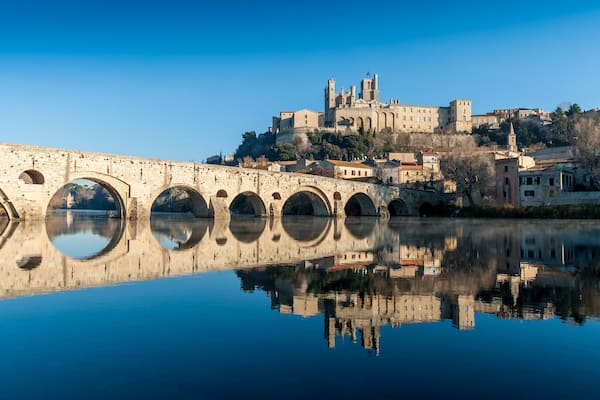 Un pont de pierre aux arches se reflétant dans l’eau calme, un bâtiment historique avec des tours en arrière-plan et un ciel bleu dégagé.