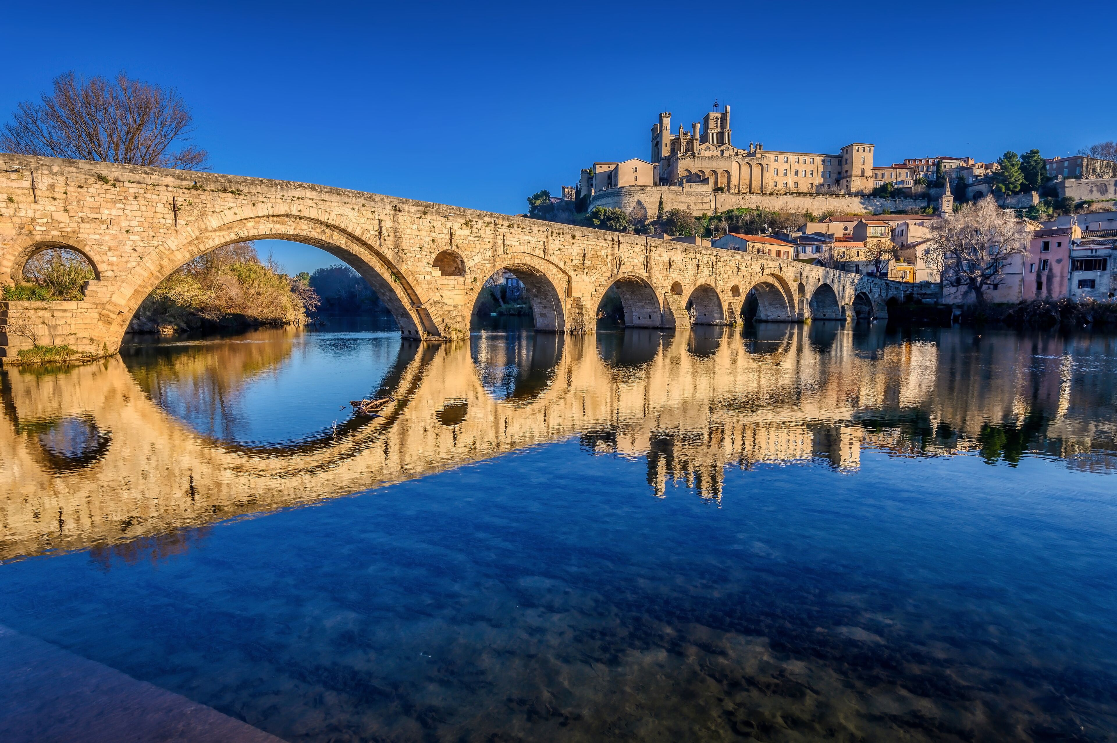 A stone bridge over a calm river with a historic building in the background.