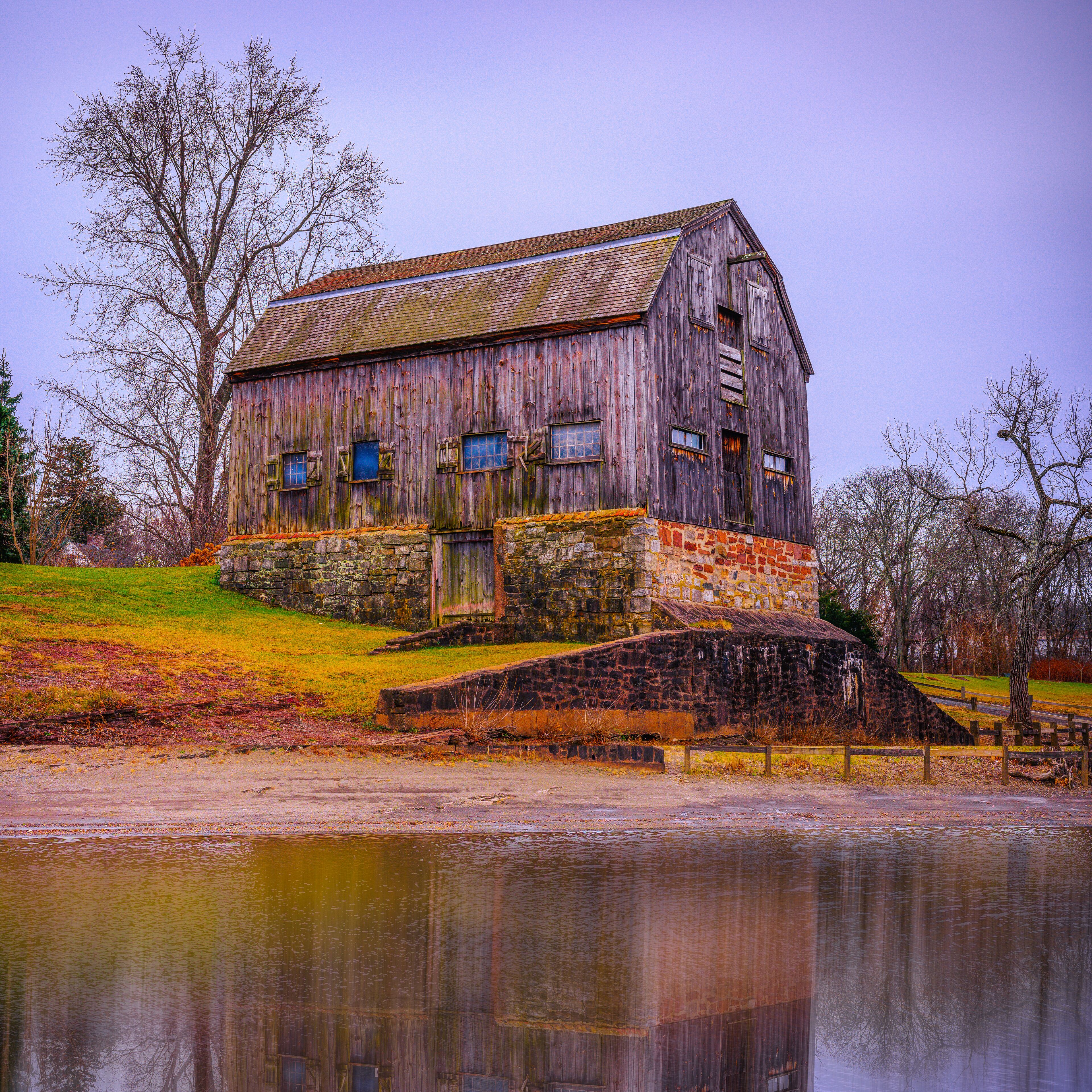 A rustic wooden barn with a stone foundation, situated by a calm body of water, surrounded by trees and grassy fields.