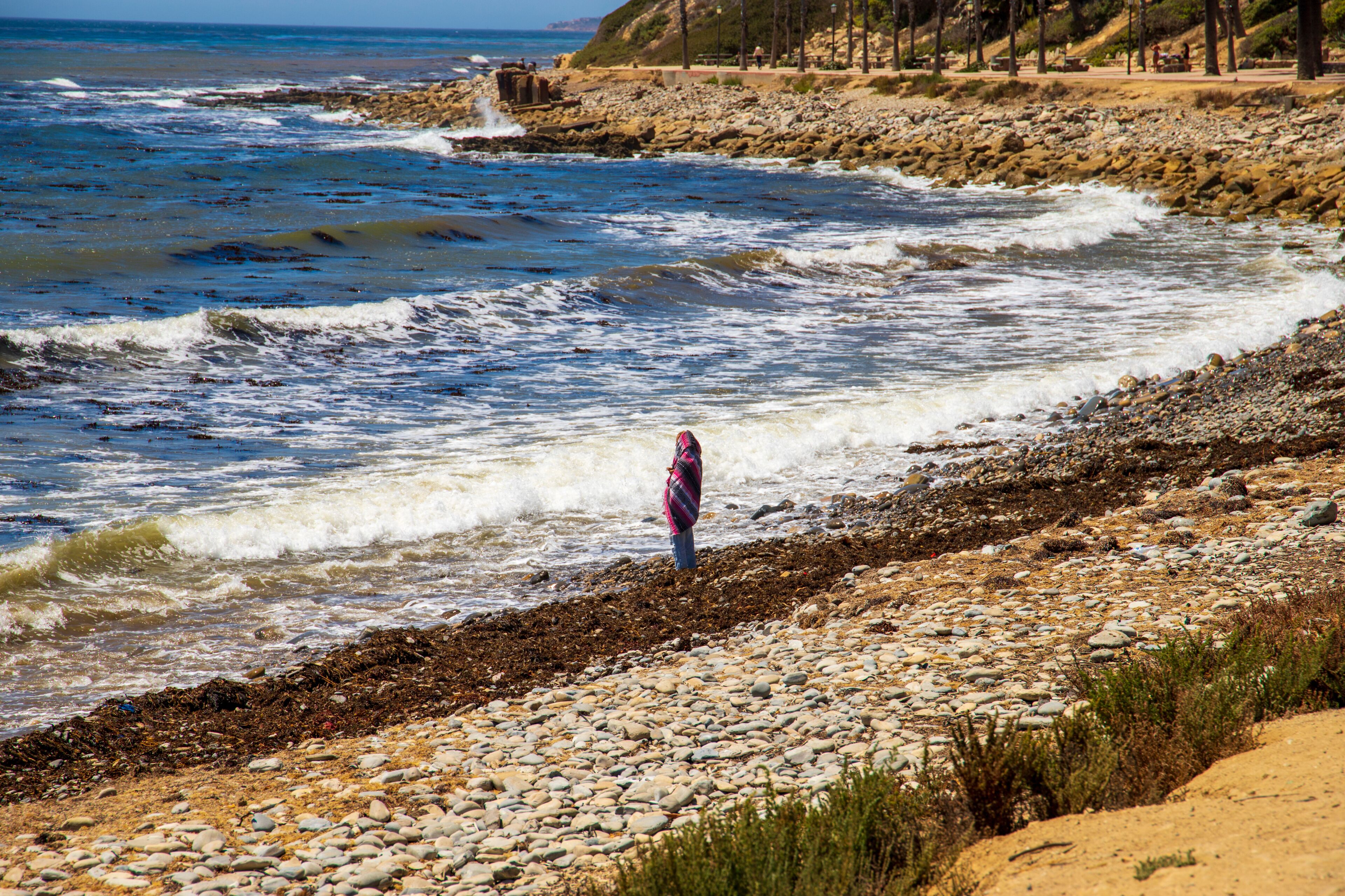 A rocky shoreline with waves crashing onto the beach, a person standing in the water, and a distant view of a rocky cliff with trees.