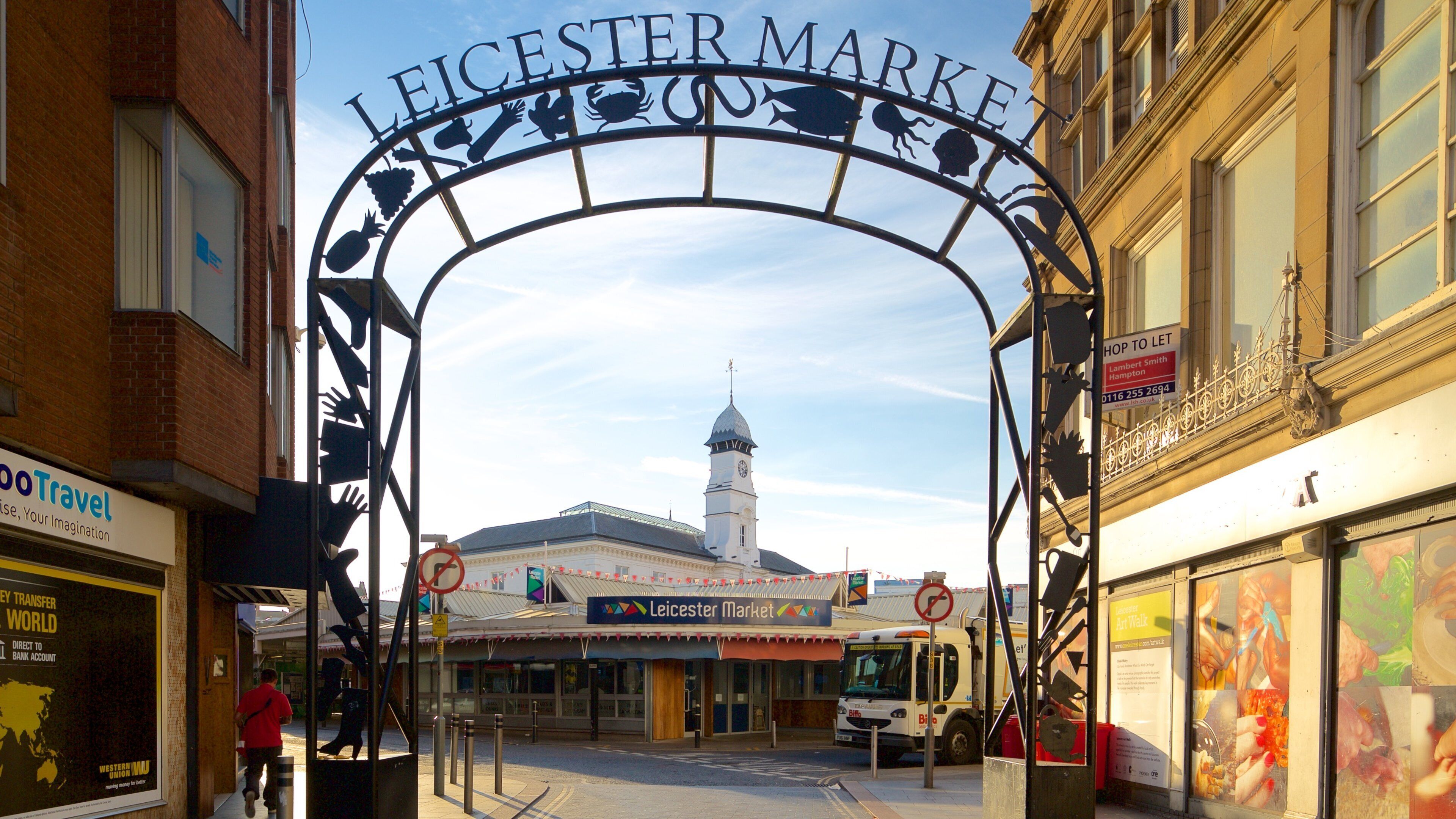 The entrance to Leicester Market with a prominent archway, a clock tower, and various shops and stalls.