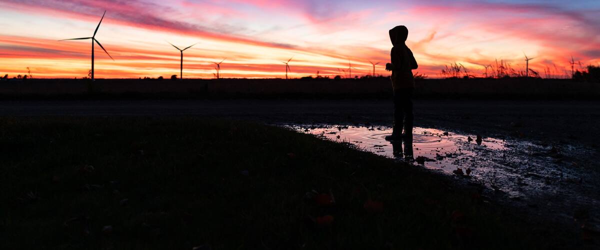A silhouette of a person standing in a field with wind turbines in the background during sunset.