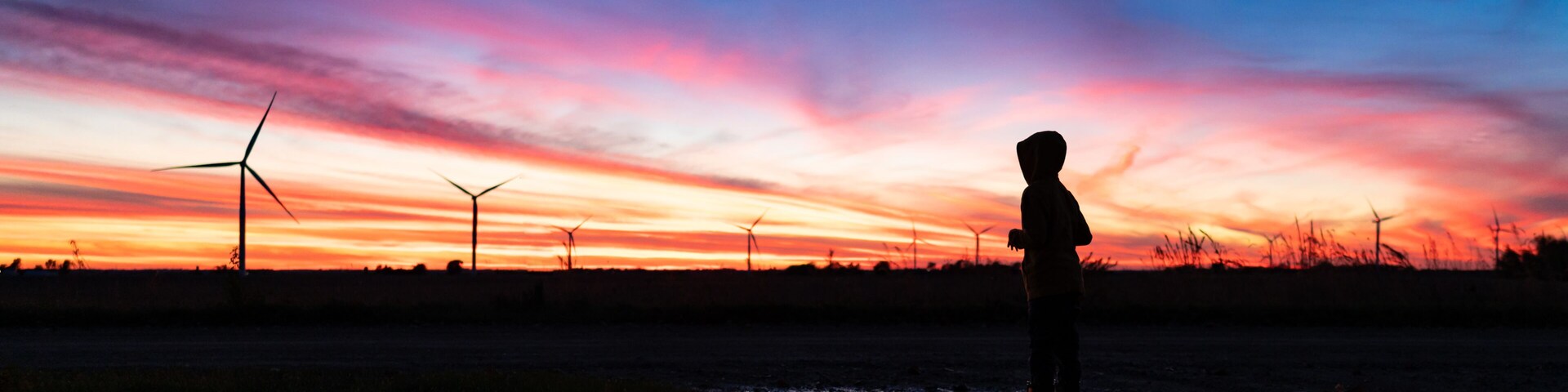 A silhouette of a person standing in a field with wind turbines in the background during sunset.