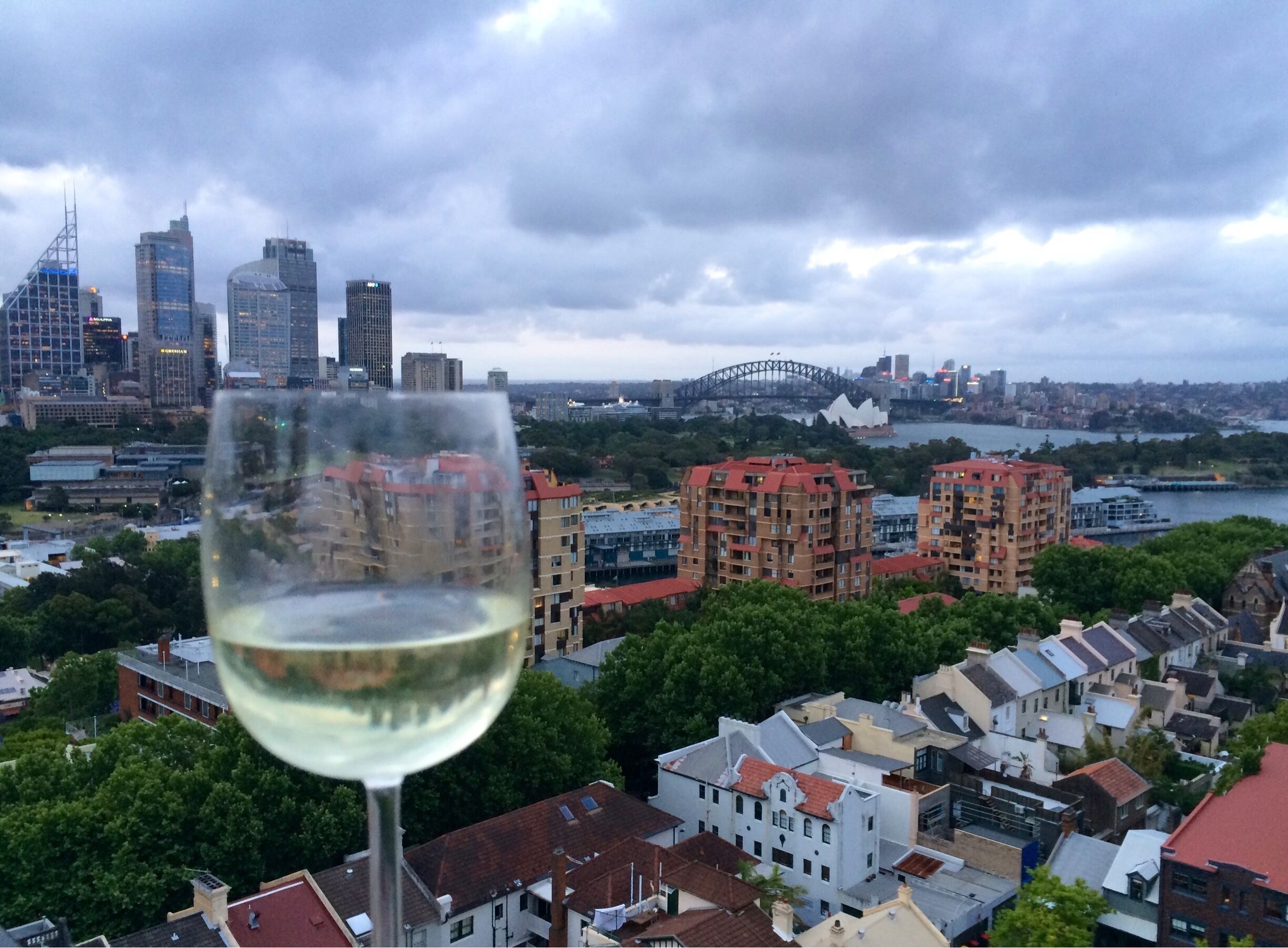 A glass of white wine with a cityscape in the background, featuring a mix of modern and traditional buildings, a bridge, and a waterfront.