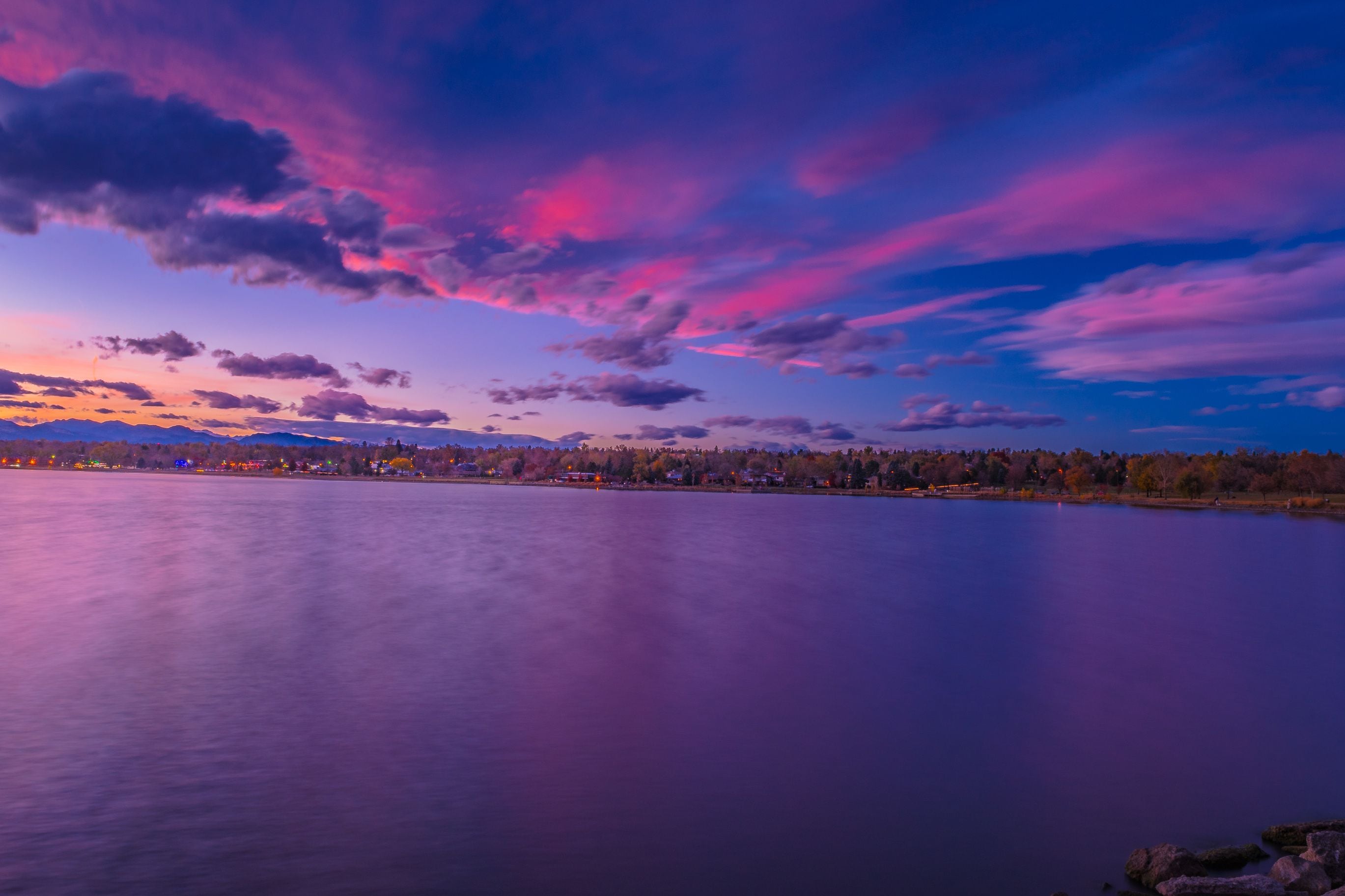 A calm lake with a cityscape in the distance, under a vibrant sky with pink and purple clouds.