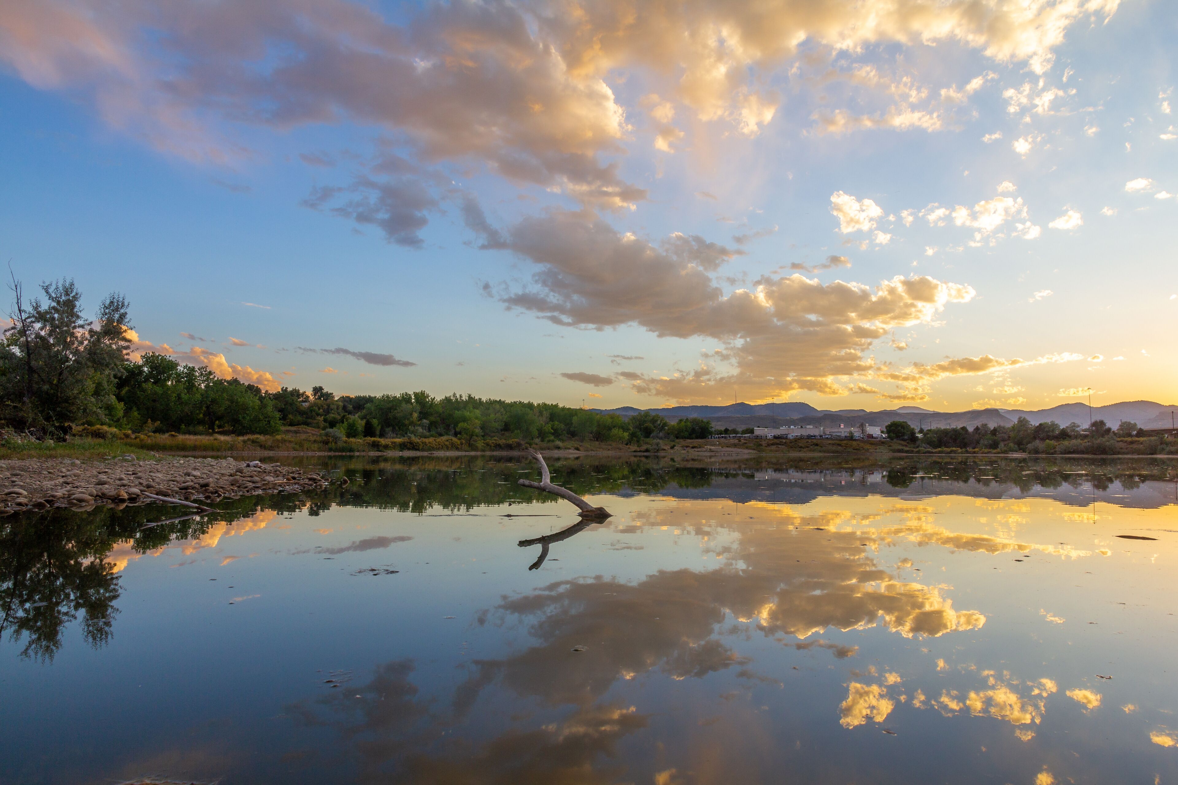 A calm river reflecting the sky with clouds, surrounded by greenery and distant mountains.