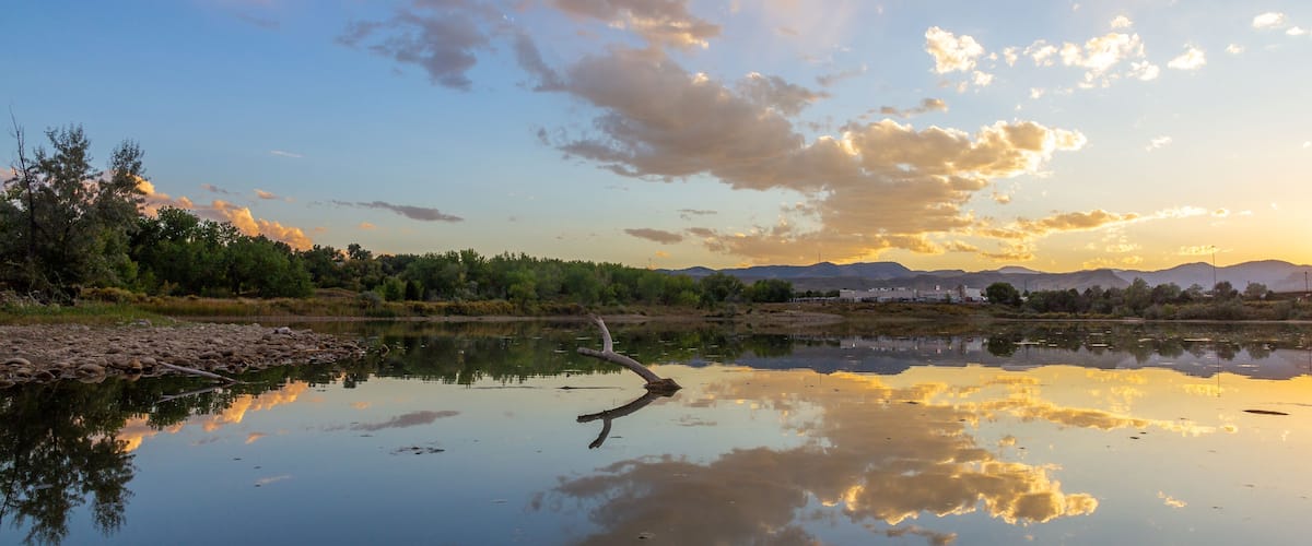 A calm river reflecting the sky with clouds, surrounded by greenery and distant mountains.