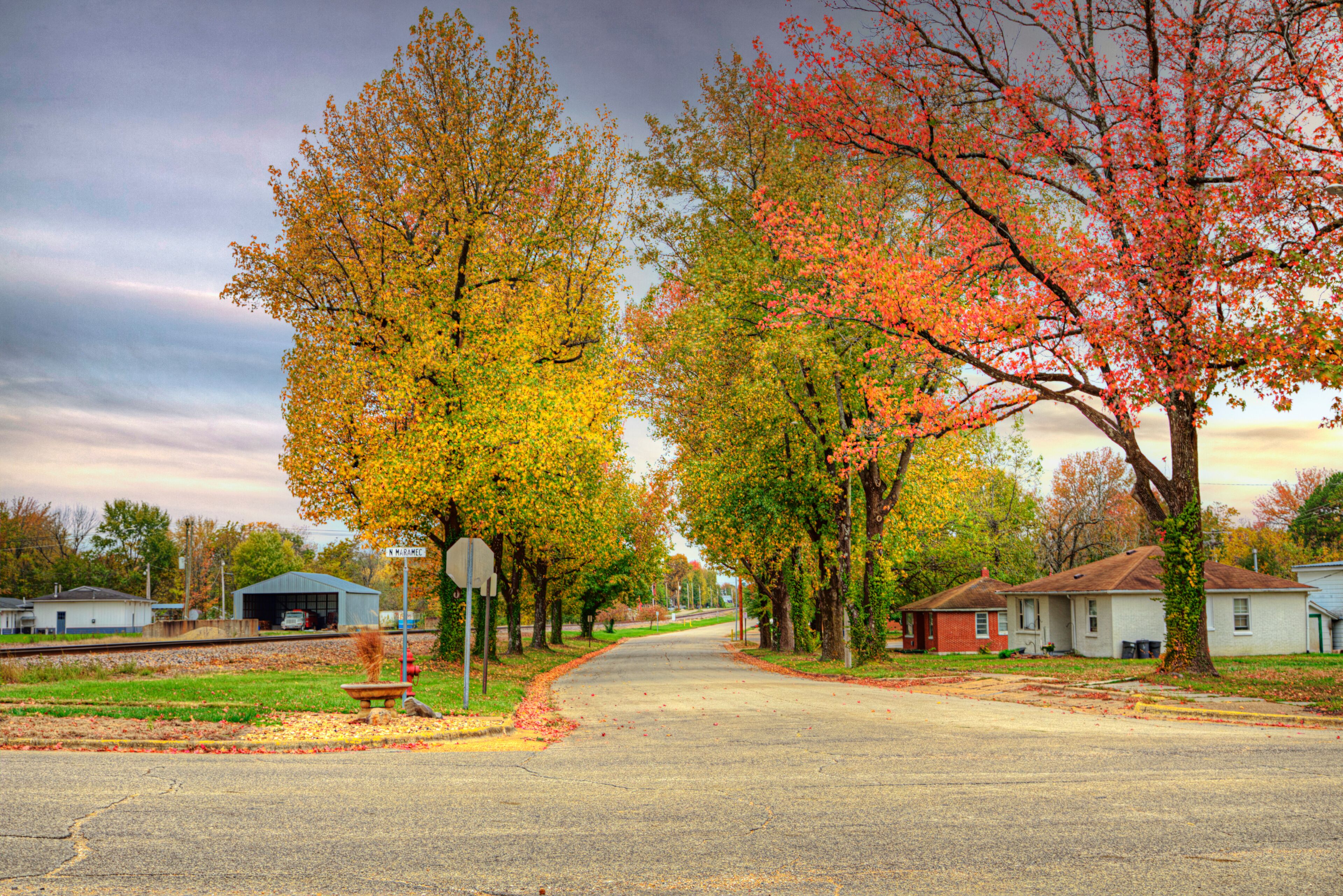A suburban street lined with autumn-colored trees, a railroad crossing, and residential houses.