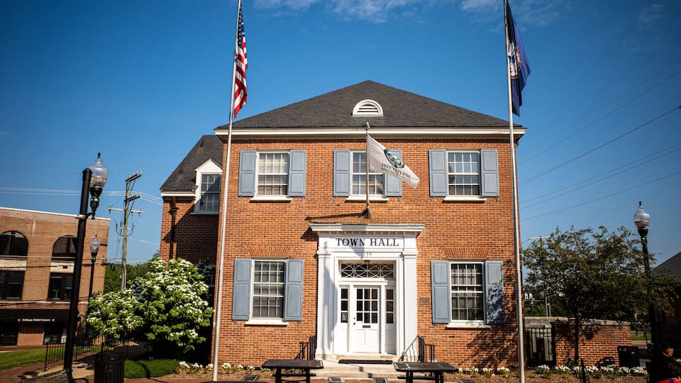 A brick town hall with a white door, blue shutters, and two flags.