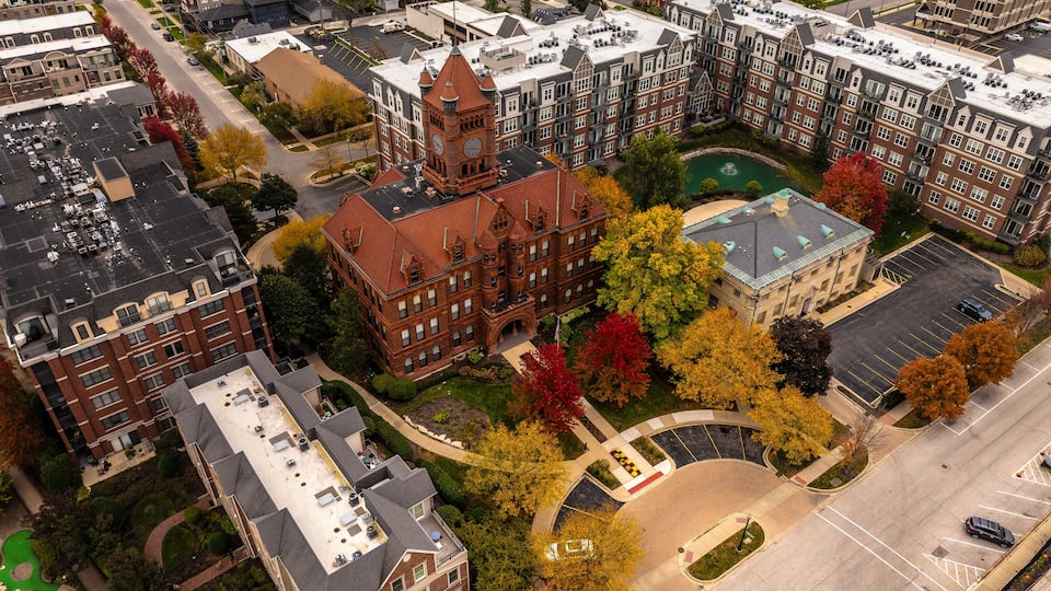 An aerial view of a residential area with a prominent red brick building with a clock tower, surrounded by trees with autumn foliage, and a parking lot.