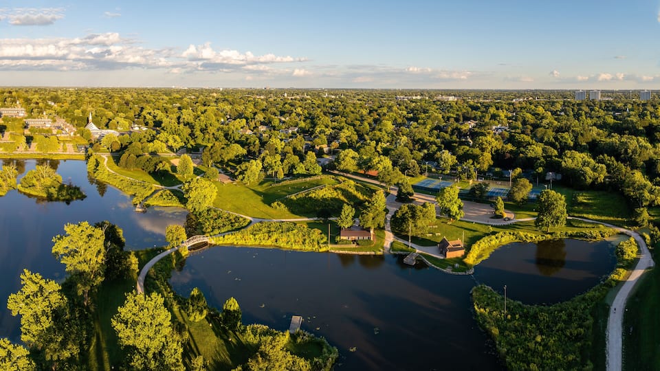 An aerial view of a park with a lake, walking paths, tennis courts, and surrounding greenery.