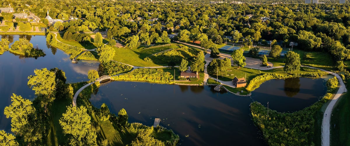 An aerial view of a park with a lake, walking paths, tennis courts, and surrounding greenery.