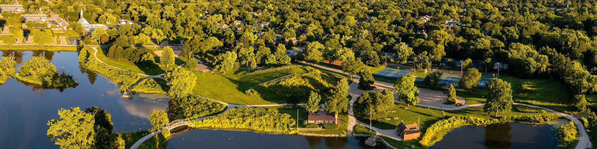 An aerial view of a park with a lake, walking paths, tennis courts, and surrounding greenery.
