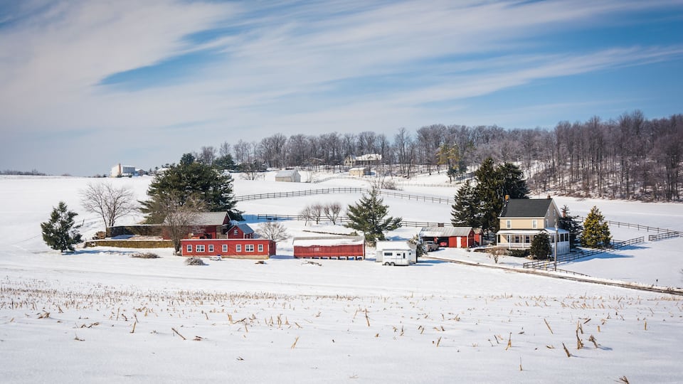 A snow-covered rural landscape with a barn, a house, and a fenced field.